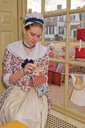 a young Colonial woman sitting at a window quilting