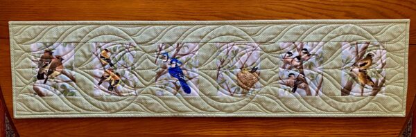 cotton quilted table runner with six colourful images of birds on a sage green background with white pin dots