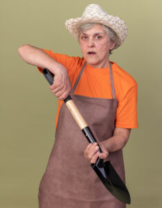 Grandmother dressed in an orange t-shirt and medium brown overalls wearing a straw hat and holding a shovel in her hands
