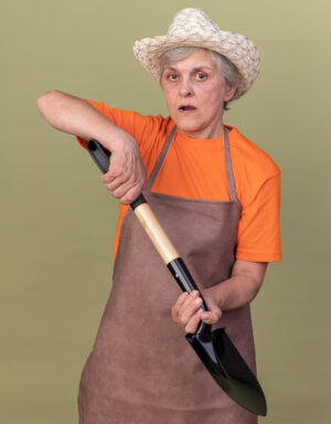 Grandmother dressed in an orange t-shirt and medium brown overalls wearing a straw hat and holding a shovel in her hands