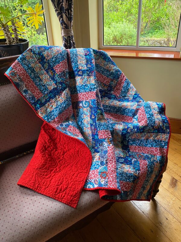 A colourful throw quilt in blues, reds and a mix of colours and part of the back showing thrown over a sofa