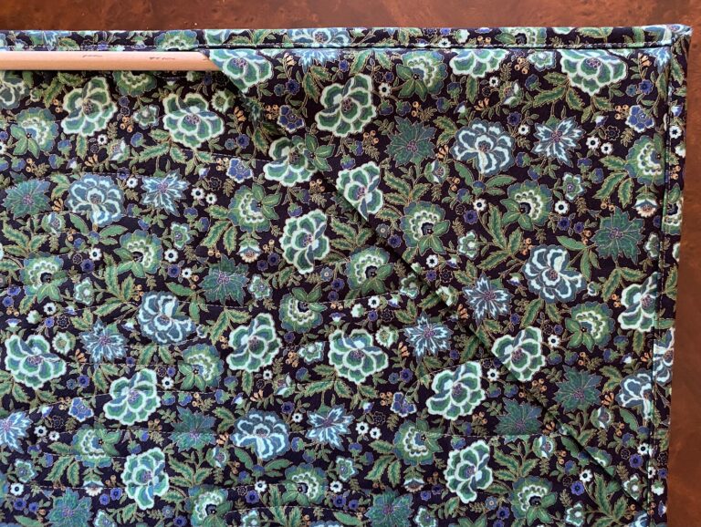 backing fabric of blue and green flowers and leaves with a hanging corner with a wooden dowel on the back of a wall hanging