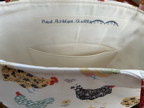 Open fabric tote with a colorful hen print; white interior lining and a stitched label reading 'Red Bridge Quilts', plus a small inner pocket.