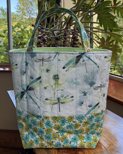 a quilted cotton tote bag featuring dragonflies and daisies in turquoise and light green. Has an interior pocket.