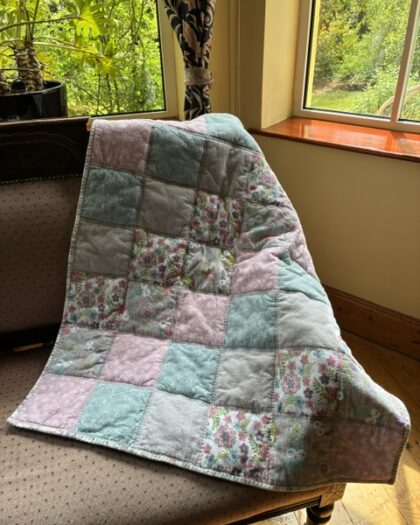 Patchwork quilt in pastel pink, blue, and floral squares draped over a chair by a sunlit window.