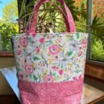 Floral fabric tote bag with pink bottom and pink handles on a wooden table by a window.