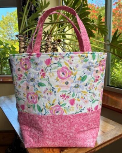 Floral fabric tote bag with pink bottom and pink handles on a wooden table by a window.
