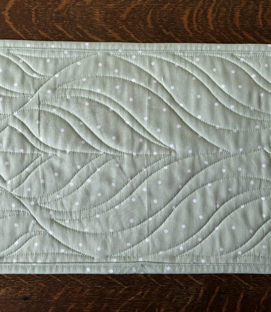 Light green quilted fabric with white polka dots and wavy leaf stitching laid on a wooden surface.