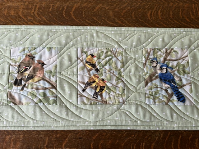 Decorative quilt border with three framed bird panels: brownish pair on the left, a yellow bird in the center, and a blue jay on the right on a pale green background.