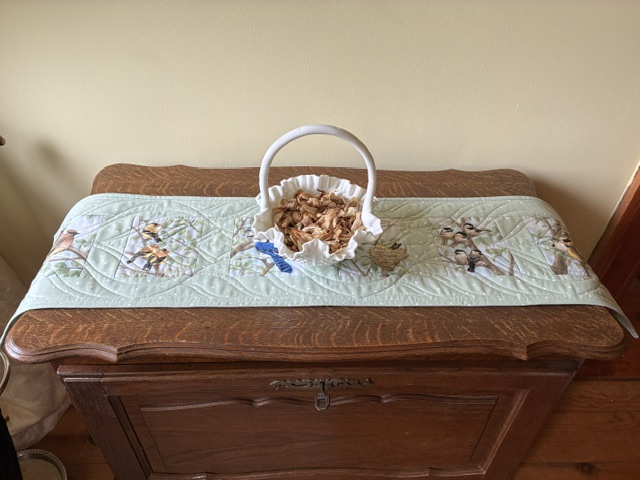 Wooden dresser with a light blue quilted runner and a white basket in the center filled with dried leaves/post-dried petals.