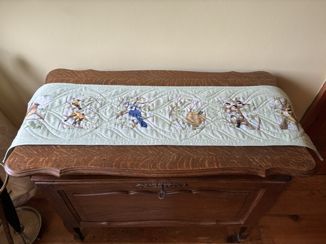 Light blue quilted table runner with colorful bird appliqués laid across an antique wooden sideboard/console table