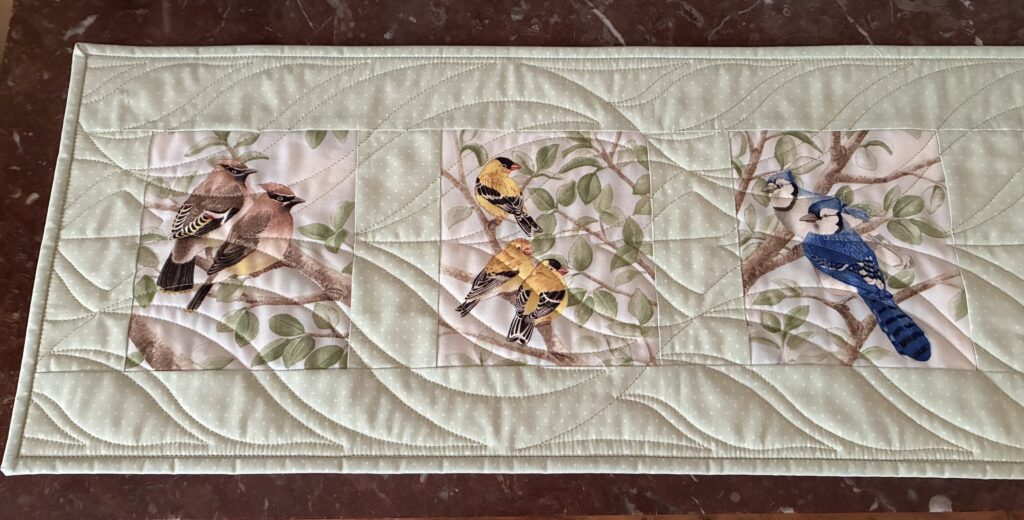 Quilt panel featuring colorful birds perched on branches with green leaves on a pale background.