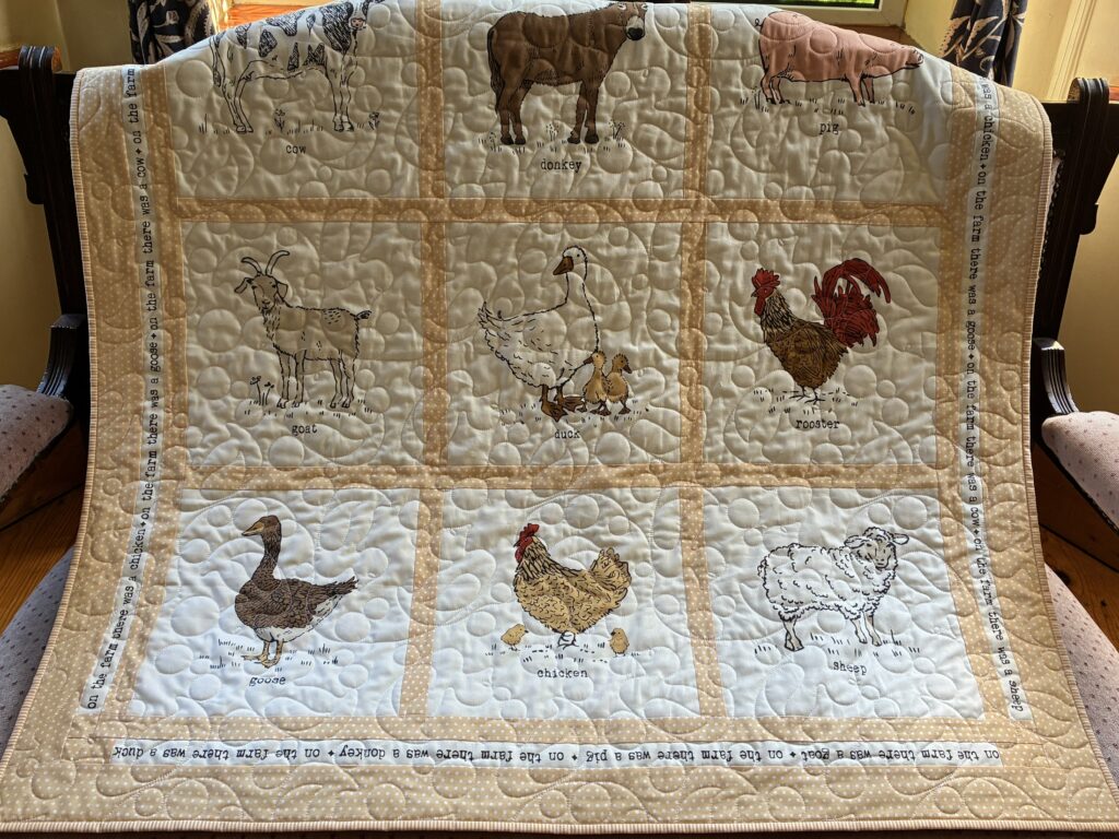 Quilted farm-themed blanket draped over a chair, showing animals labeled with names: cow, donkey, pig, goose, duck, chicken, rooster, sheep, goat.