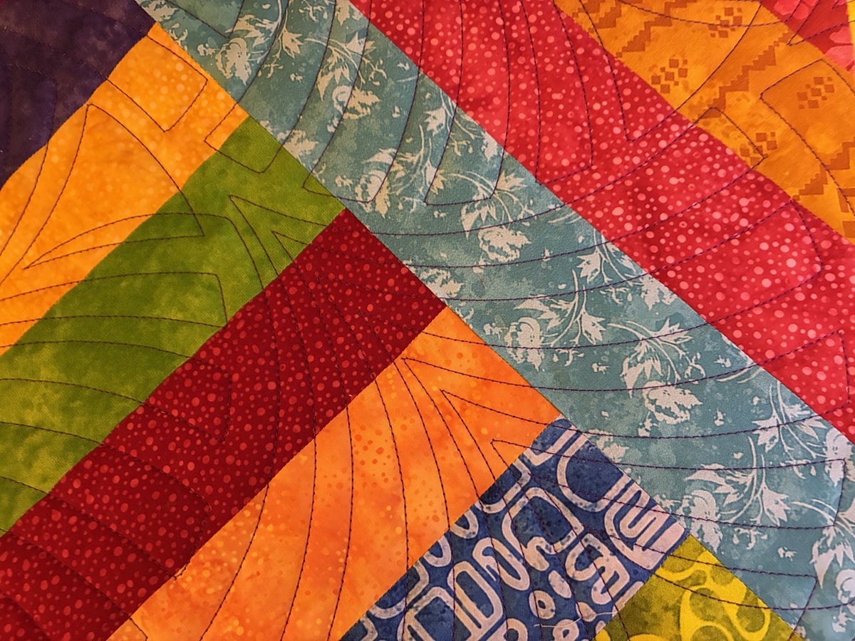 Very scrappy multi-coloured quilt with diagonal strips quilted with a fan-like design in purple thread